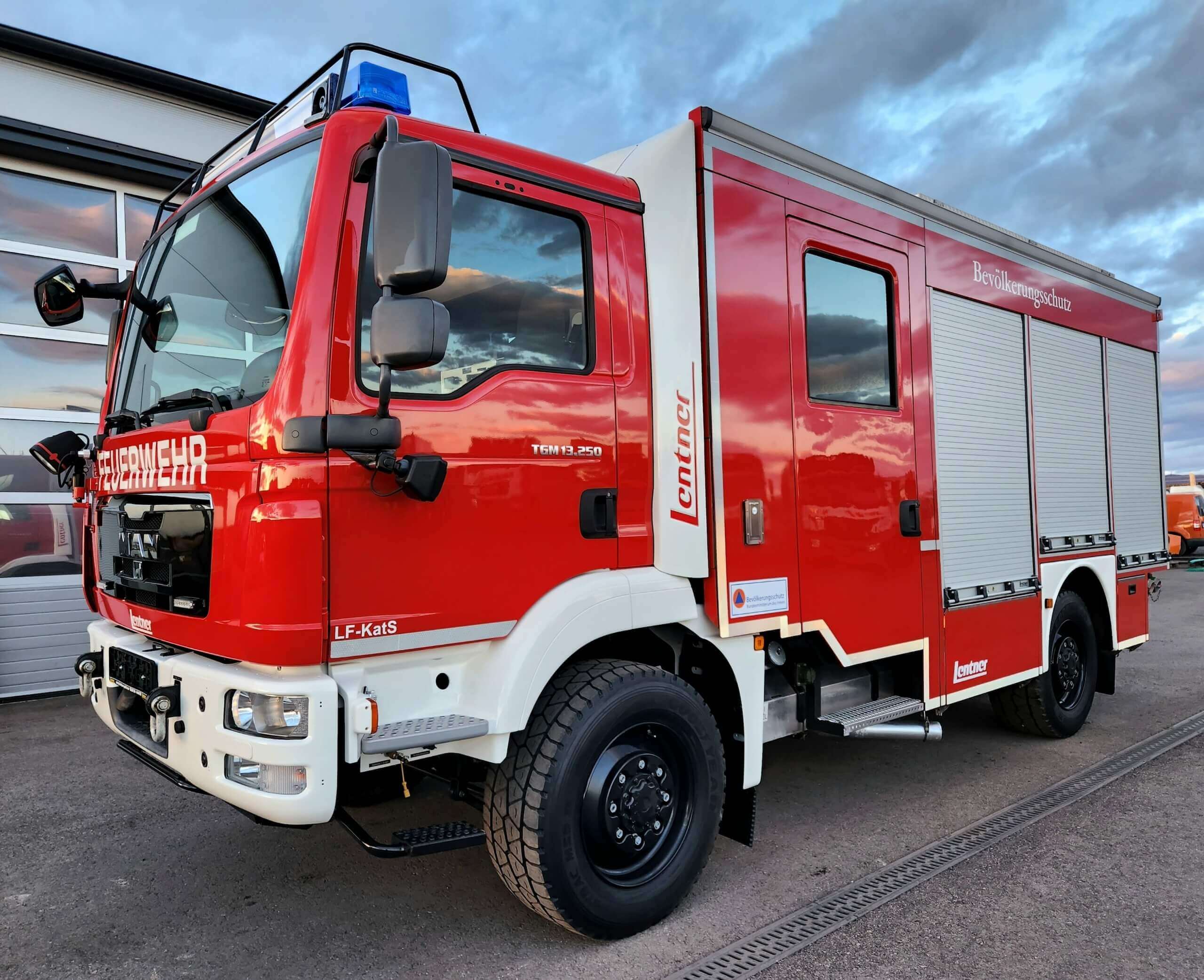 Rotes Feuerwehrauto mit blauen Lichtern vor einem Gebäude bei Sonnenuntergang. Feuerwehrfahrzeug von MAN.