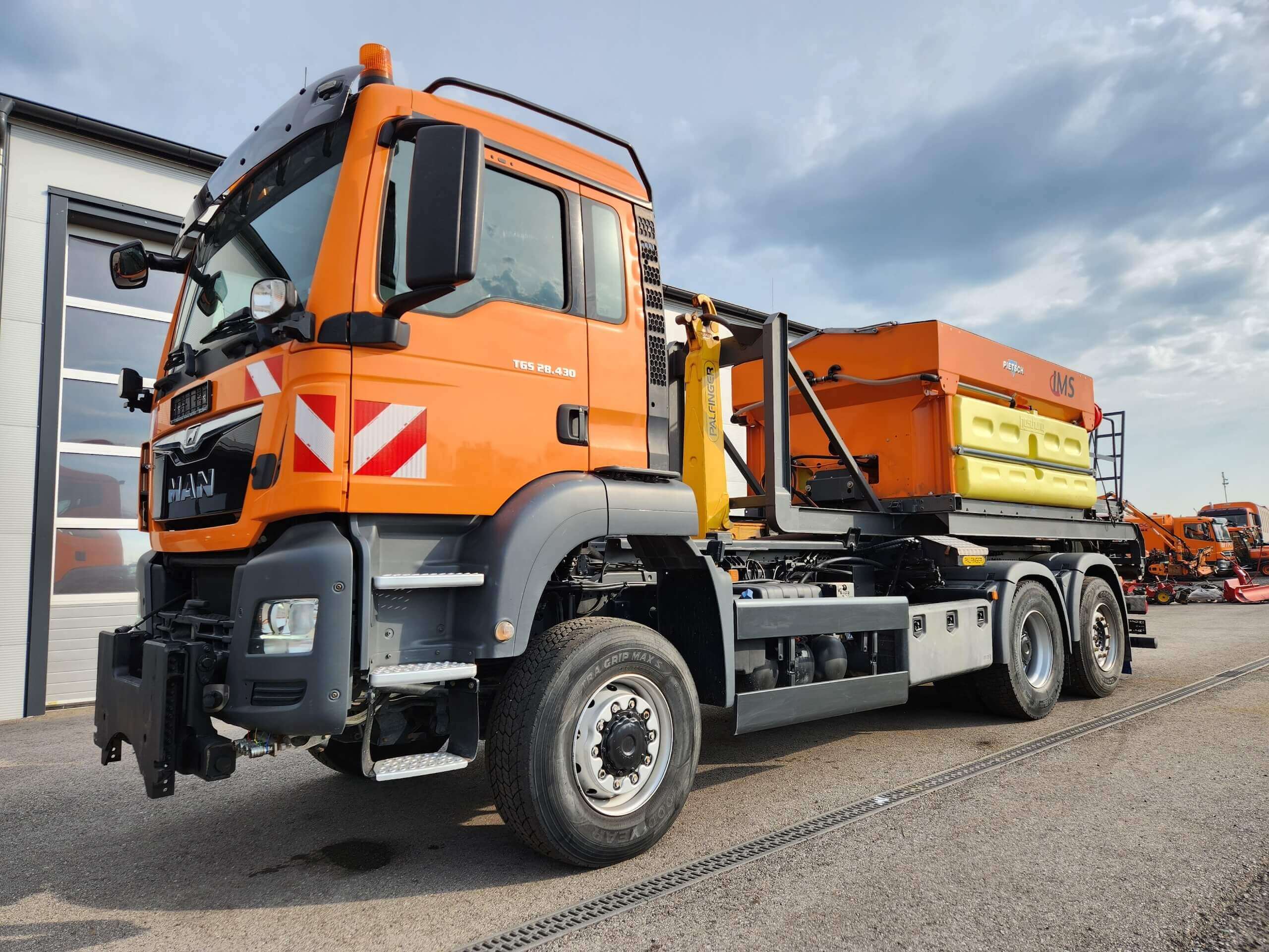 Orange MAN LKW mit Streugerät für Winterdienst auf Betriebshof bei bewölktem Himmel.