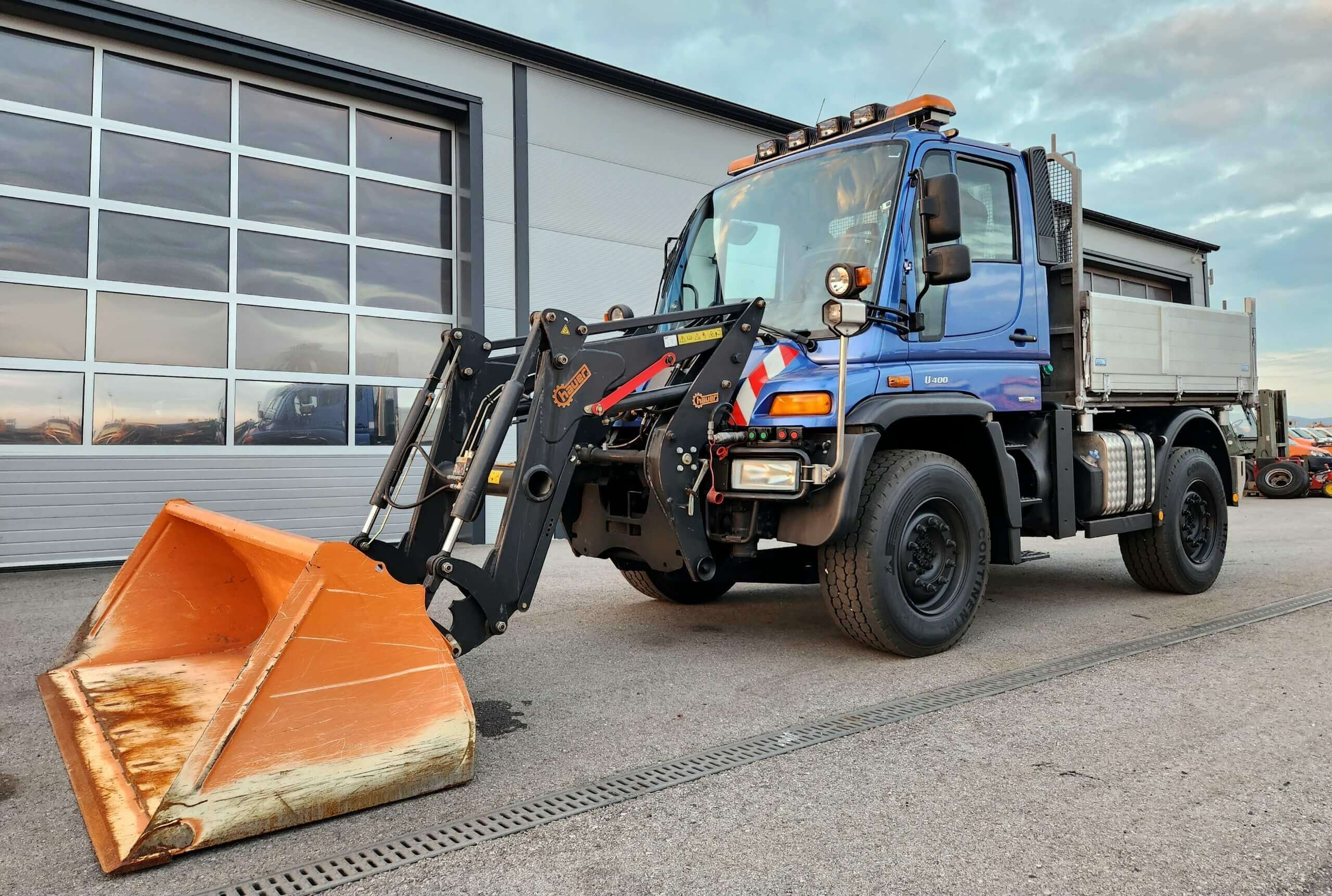 Blauer Unimog U 400 mit orangem Schneeschild auf einem Betriebshof vor großen Garagentoren abgestellt.